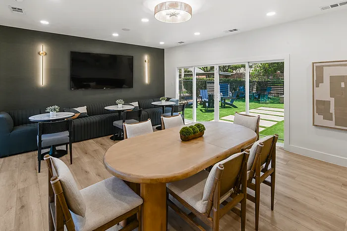 Chic clubhouse with elegant wooden dining table, cozy seating, large TV, and garden views through sliding glass doors.