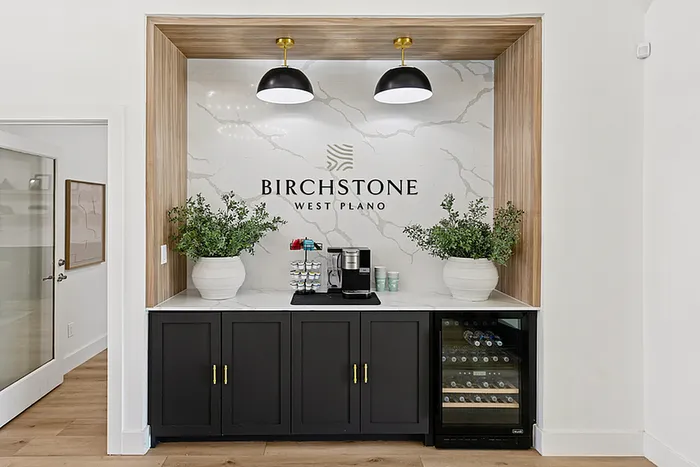 Sophisticated coffee bar at Birchstone West Plano featuring elegant cabinetry, modern lighting, and a built-in wine cooler with lush greenery for a welcoming touch.