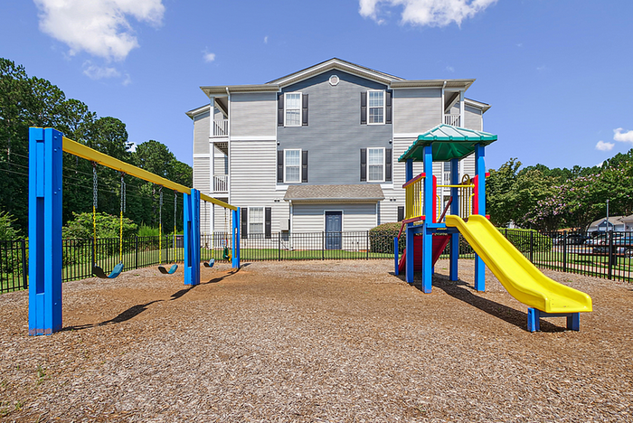 Inviting apartment complex with a vibrant playground, featuring swings and a colorful slide, set against a backdrop of lush greenery and clear blue skies.