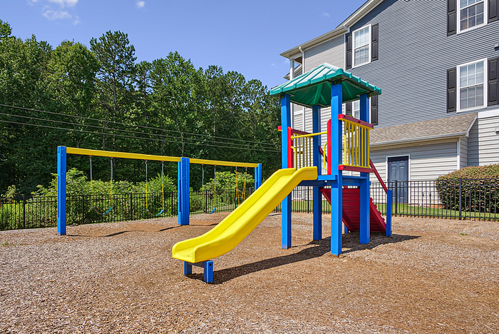 Charming playground area featuring a colorful slide and swings, nestled next to a modern apartment building with lush greenery in the background.
