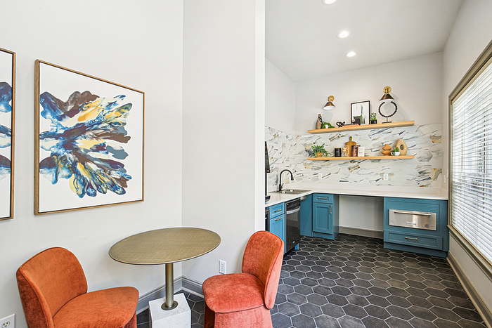 Chic apartment space showcasing a stylish nook with vibrant orange chairs, modern art, and a sleek kitchen corner with blue cabinetry and open shelving.