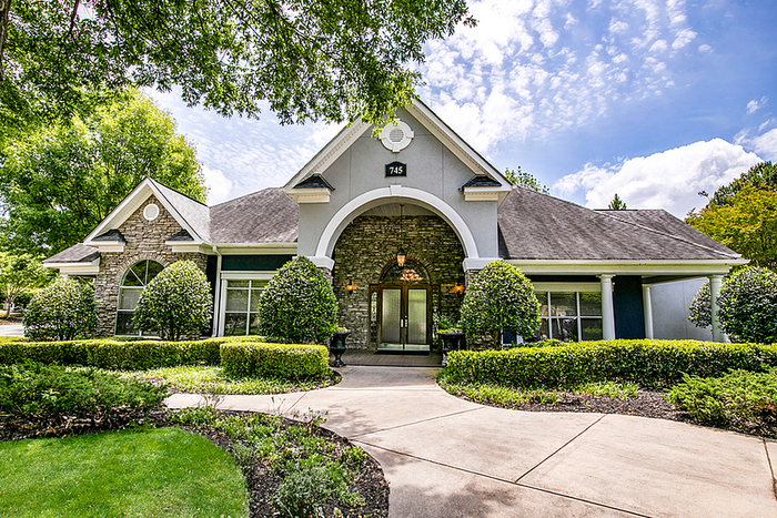 Charming stone and stucco house with lush green landscaping and a circular driveway entrance, under a vibrant blue sky with clouds.