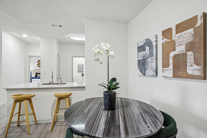 Modern apartment interior featuring a sleek kitchen breakfast bar with stylish stools, adjacent to a chic round dining table adorned with a fresh orchid, complemented by contemporary wall art.