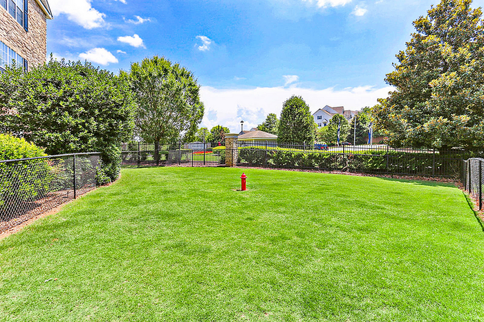 Fenced community dog park with a large, well-maintained grassy play area, mature trees, and a pet waste station, offering plenty of space for residents and their dogs to enjoy the outdoors.