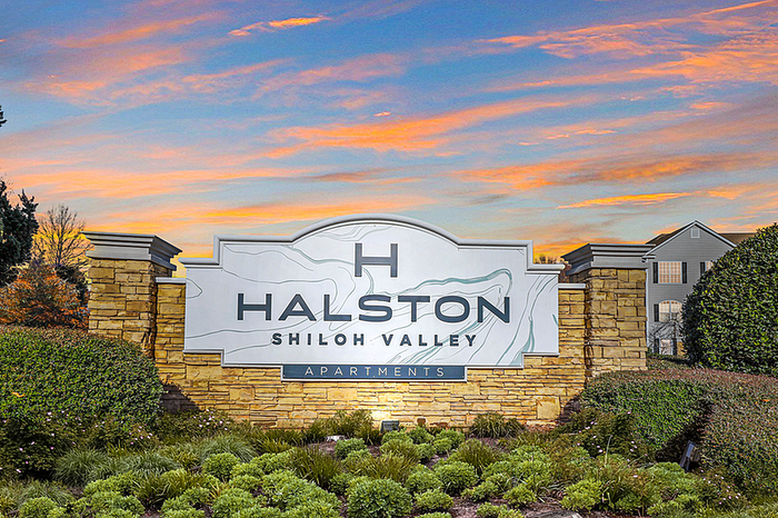 Elegant entrance sign for Halston Shiloh Valley Apartments, framed by lush greenery and a stunning sunset backdrop.