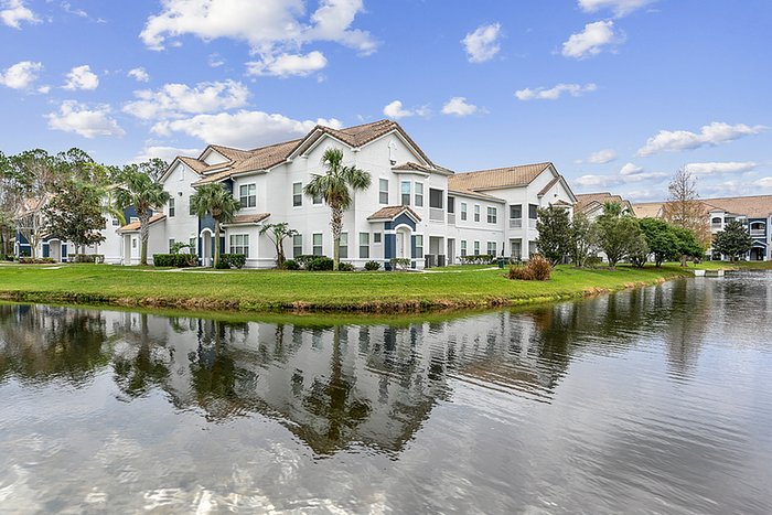 Charming community surrounded by lush greenery and a serene pond, under a bright blue, cloud-dotted sky.