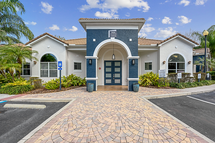 Charming entrance to a clubhouse featuring elegant architecture, manicured landscaping, and inviting paver pathways under a clear blue sky.