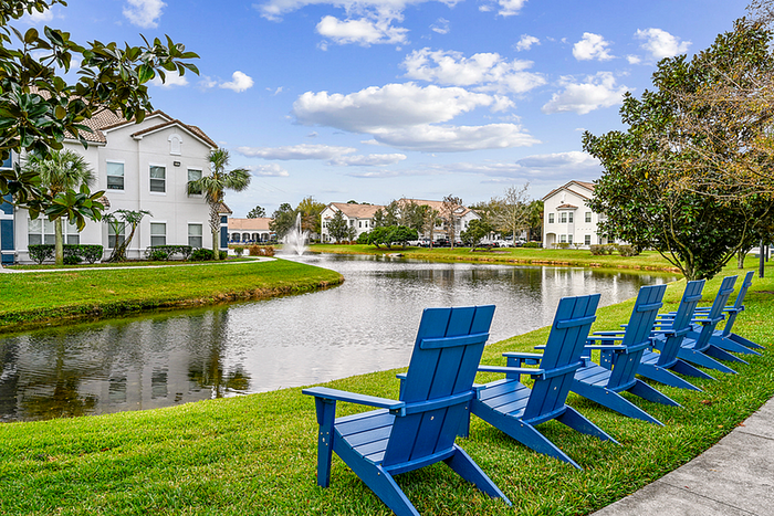 Charming apartment community with serene lake views, surrounded by lush greenery and lined with elegant blue Adirondack chairs.
