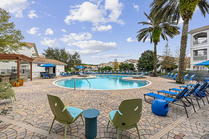Elegant apartment pool area with luxurious lounge chairs, vibrant umbrellas, and lush palm trees under a bright blue sky.