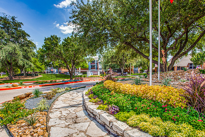 Lush apartment landscape with vibrant flowerbeds, mature trees, and a serene pathway under a clear blue sky.