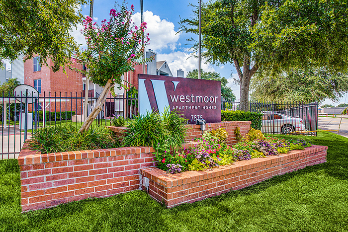 Charming entrance to Westmoor Apartment Homes featuring vibrant landscaping, red brick sign, and lush greenery.
