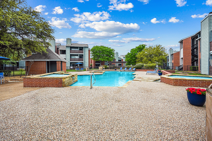 Inviting outdoor pool area with brick accents and lush landscaping, surrounded by modern apartment buildings under a vibrant blue sky.