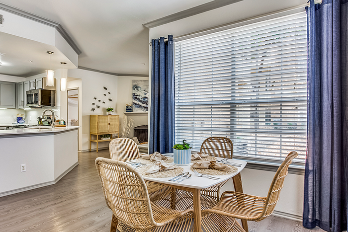 Bright and inviting dining area with wicker chairs and a modern kitchen in view, surrounded by large windows with elegant curtains.