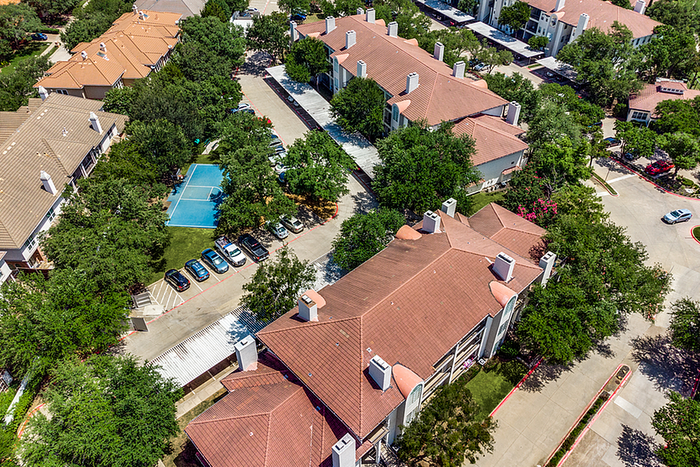 Charming apartment community with terracotta roofs, surrounded by lush greenery and featuring a central sports court and ample parking.