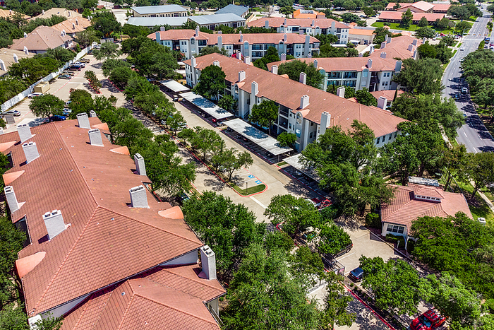 Aerial view of a charming apartment community surrounded by lush green trees, showcasing elegant red-tiled roofs and a well-maintained road connecting the community.