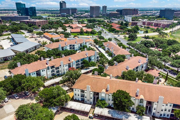 Aerial view of charming apartment community with terracotta roofs amid lush greenery, set against a backdrop of modern city skyscrapers and clear blue sky.