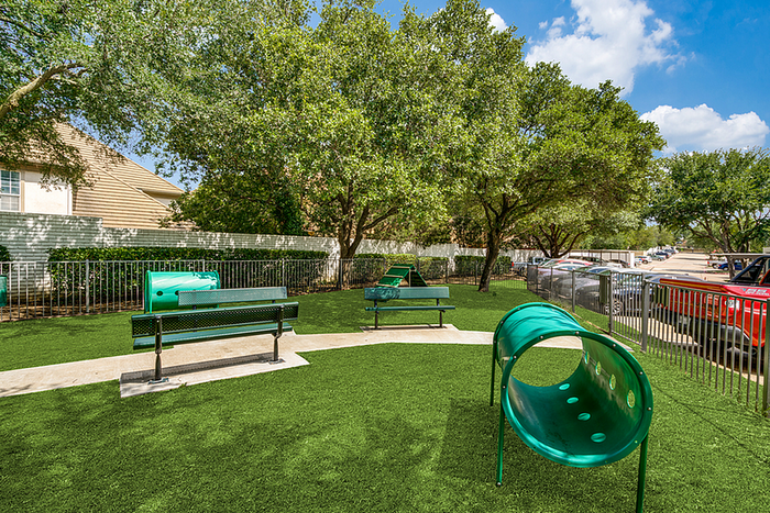 Vibrant apartment community dog park featuring lush green turf, benches, and playground tunnels under a canopy of trees.