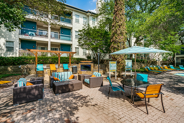 Charming outdoor patio area with stylish seating under vibrant umbrellas, surrounded by lush greenery and modern apartment buildings.