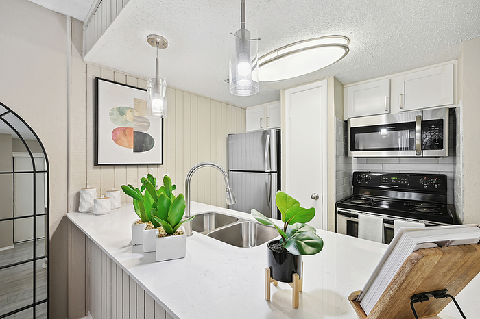 Modern kitchen with sleek stainless steel appliances, stylish pendant lighting, and elegant white cabinetry, accented by vibrant green plants and artistic wall decor.
