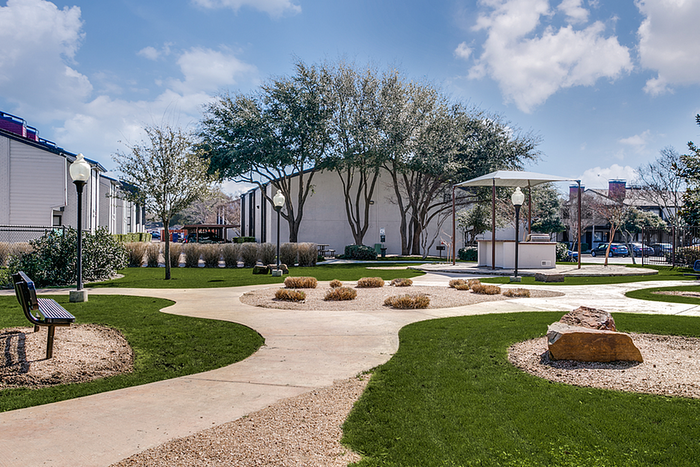 Charming apartment community with well-manicured green spaces, shaded seating areas, and picturesque walking paths under a vibrant blue sky.