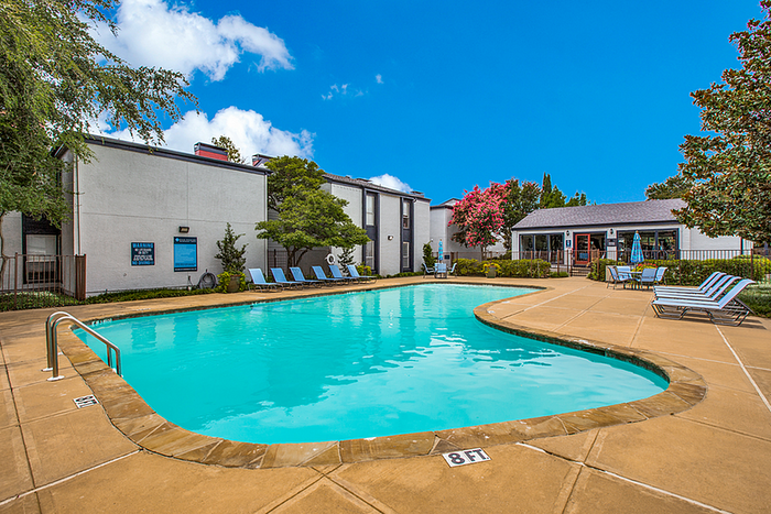 Inviting apartment pool area with crystal-clear water, surrounded by comfortable lounge chairs and lush landscaping under a bright blue sky.