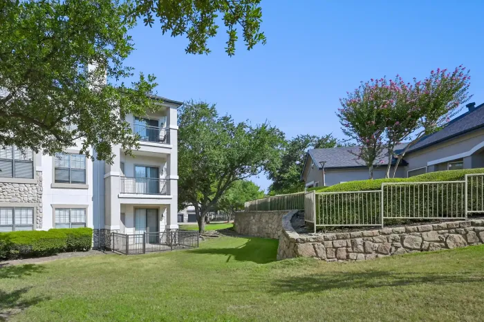 Landscaped courtyard between apartment buildings featuring mature trees, green lawn, private balconies, and stone retaining walls.
