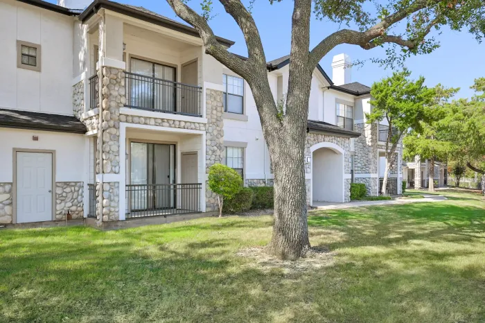 Exterior of an apartment building featuring private balconies, stone accents, mature trees, and a landscaped lawn.
