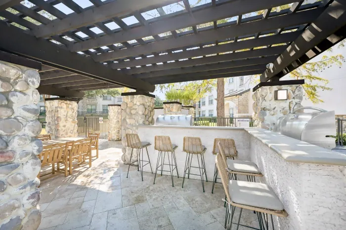 Shaded outdoor grilling and dining area with bar seating beneath a pergola near the community pool.