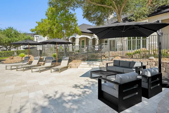 Poolside lounge area with shaded seating, lounge chairs, and umbrellas surrounded by landscaped grounds.