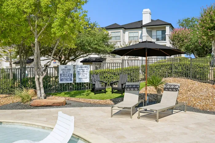 Poolside lounge chairs with a shaded umbrella and landscaped seating area surrounded by apartment homes.