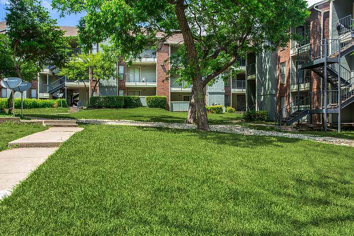 Inviting apartment complex with lush green lawns, mature trees, and modern staircases under a bright blue sky.