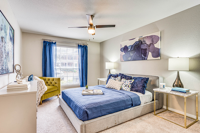 Elegant bedroom with a cozy atmosphere, featuring a plush blue bedspread, modern art, and natural light through wide windows with blue curtains.