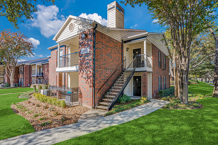 Charming red-brick apartment building with lush green landscaping and a cozy balcony under a clear blue sky.