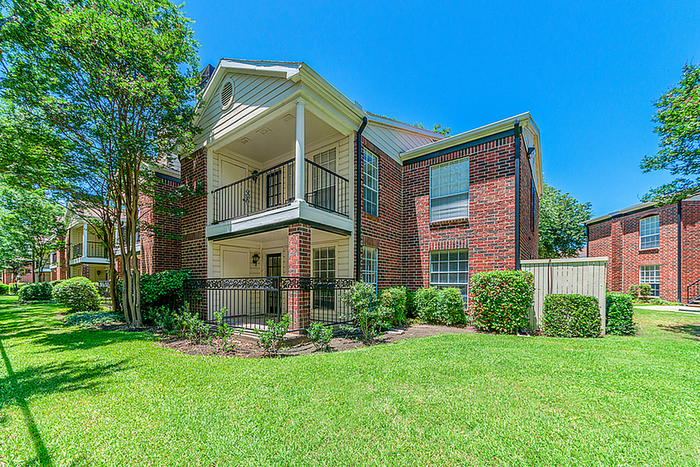 Charming red brick apartment building with private balconies, lush greenery, and vibrant lawn under a clear blue sky.