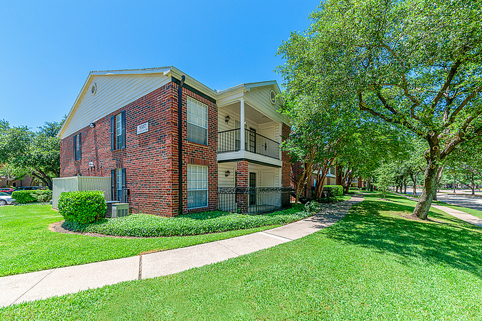 Charming two-story brick apartment with private balconies, surrounded by lush greenery and a peaceful walkway.