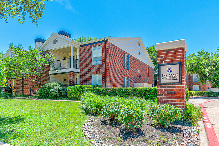 Charming brick apartment community 'The Oaks at Valley Ranch' surrounded by lush greenery and a bright blue sky.