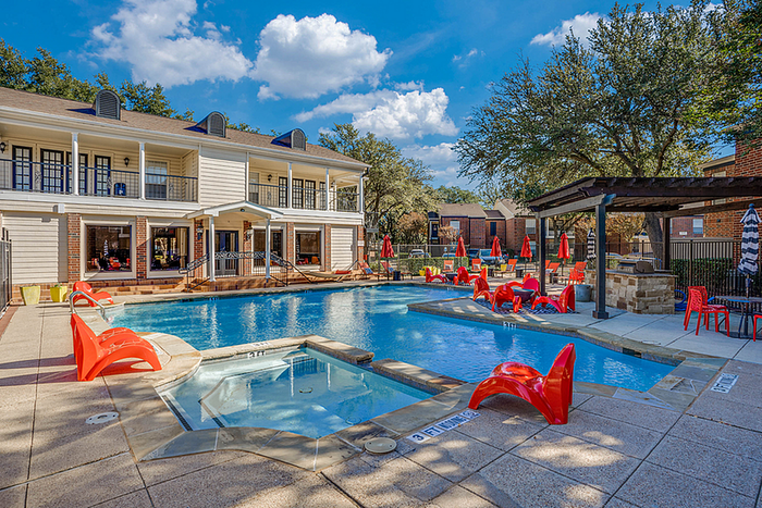 Vibrant apartment community swimming pool area with stylish red lounge chairs, a jacuzzi, and elegant seating under a charming pergola amid lush greenery.