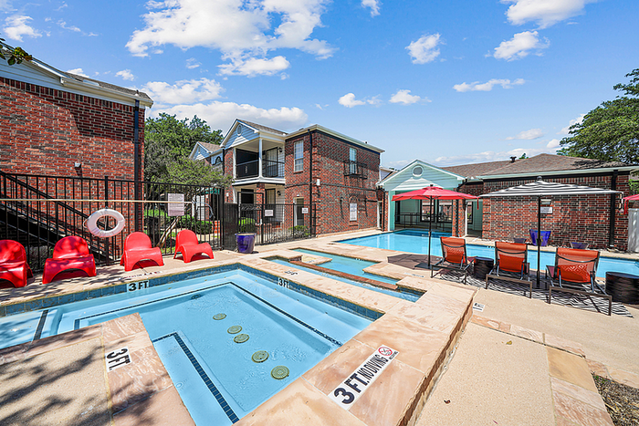 Inviting outdoor pool area with relaxing lounge chairs, vibrant red seating, and charming brick buildings under a bright blue sky.