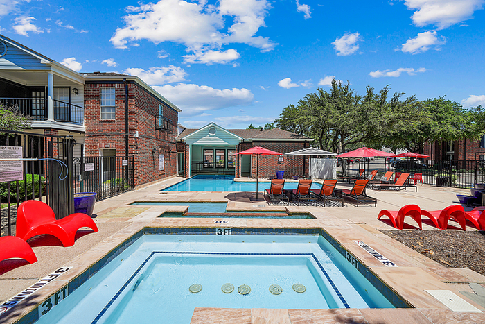Inviting apartment community with a swimming pool, vibrant red lounge chairs, and beautiful brick buildings under a clear blue sky.