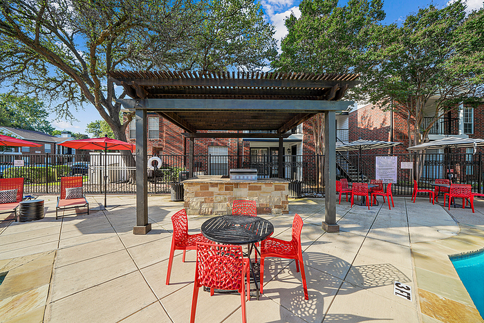 Charming outdoor patio with vibrant red seating, shaded pergola, and inviting poolside ambiance in a serene apartment complex.