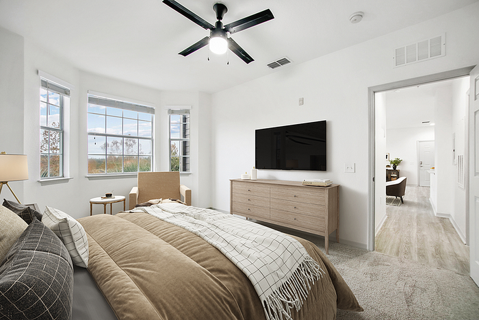 Bright and airy bedroom with large windows, cozy bed, modern ceiling fan, and sleek décor, leading into a spacious hallway.