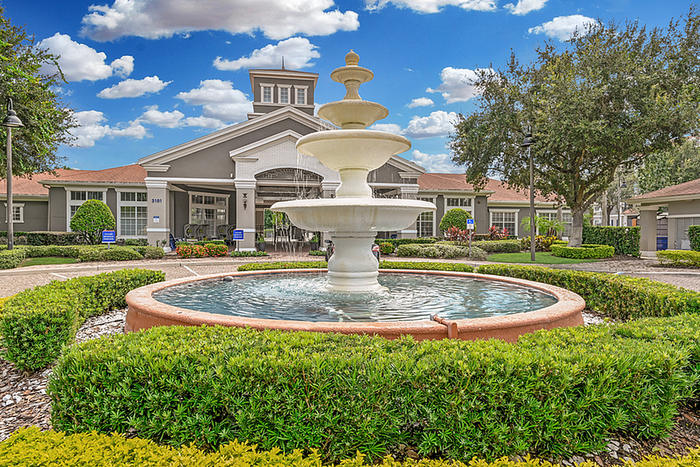 Charming apartment community entrance with a grand tiered fountain surrounded by lush greenery and a stately building under a bright blue sky.
