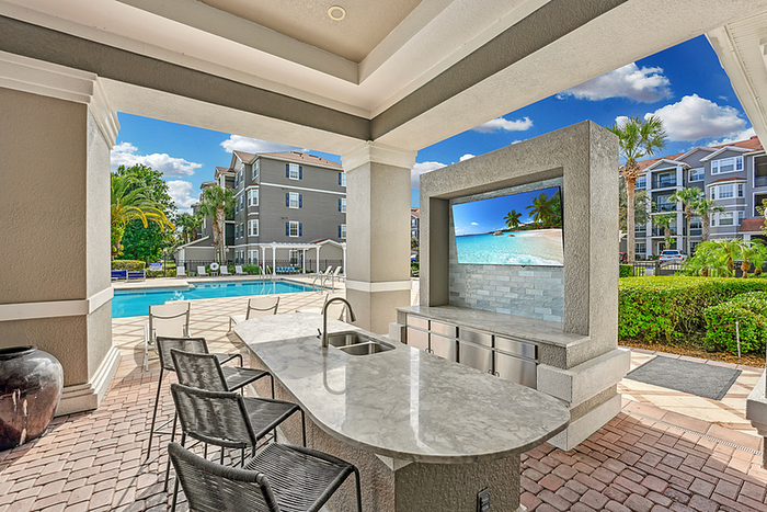 Luxurious outdoor area featuring a sleek kitchen with bar seating overlooking a sparkling pool, surrounded by lush greenery and stylish apartment buildings.