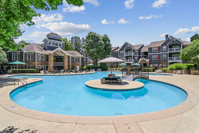 Luxurious apartment community with a sparkling swimming pool, elegant lounge chairs, and vibrant greenery under a clear blue sky.