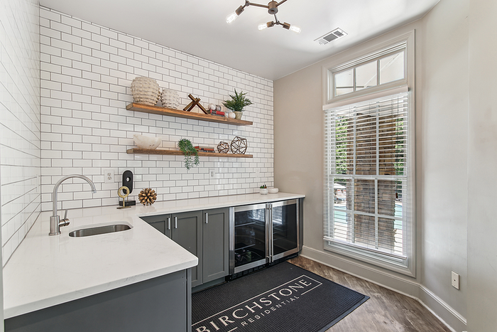 Chic clubhouse kitchenette with white subway tile backsplash, floating shelves with modern decor, and a large window overlooking a pool.