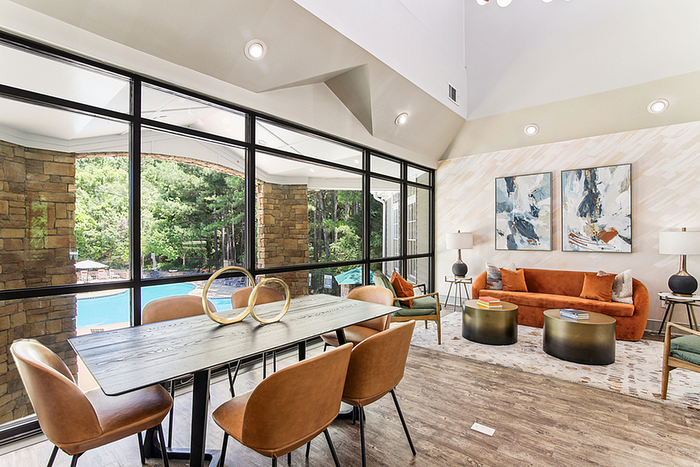 Bright and elegant clubhouse with modern chic d&eacute;cor, featuring a cozy seating area with vibrant orange sofa, stylish artwork, and expansive floor-to-ceiling windows overlooking a serene outdoor view.