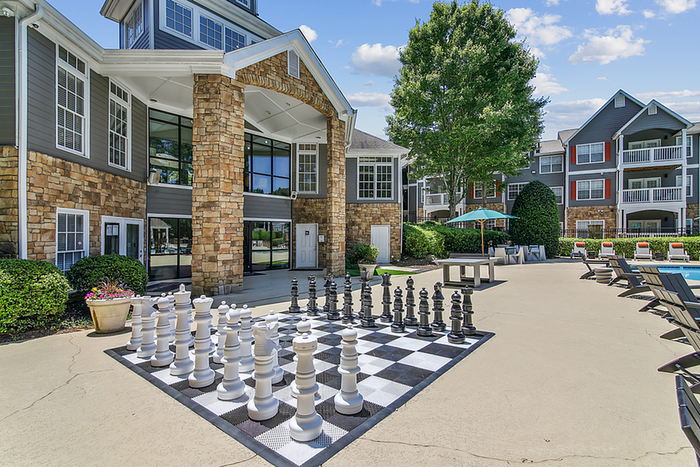 Luxurious apartment community with a grand stone facade, surrounded by lush greenery. Features an outdoor patio with a giant chess set and inviting swimming pool, perfect for relaxation and leisure.