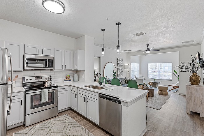 Contemporary apartment kitchen with stainless steel appliances, sleek white cabinetry, and an open-plan living room with natural light and stylish decor.