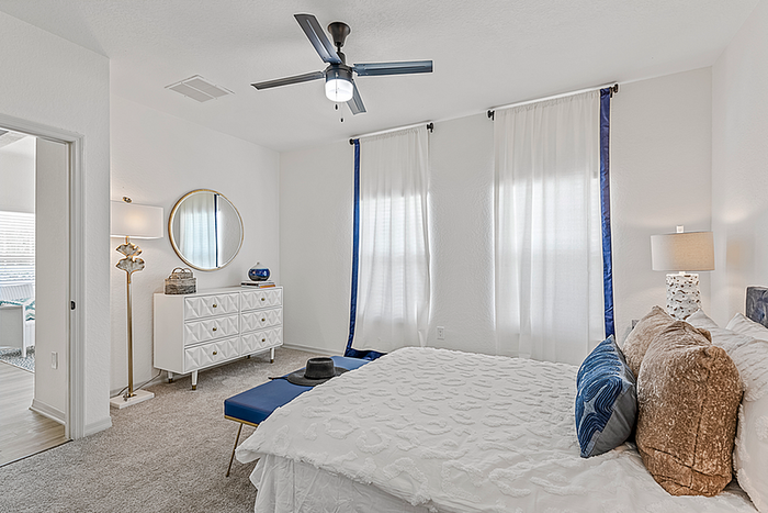 Elegant bedroom features a cozy bed with plush pillows, chic white dresser, stylish floor lamp, and a modern ceiling fan against a backdrop of bright, airy curtains.