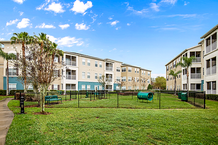 Charming apartment community with lush greenery, pet-friendly play area, and inviting patio spaces under a clear blue sky.
